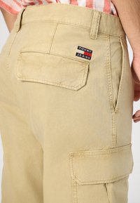 Pantaloni cargo beige con tasca a toppa sul retro e un'etichetta tessuta con "TOMMY JEANS" in rosso, bianco e blu. Texture liscia.