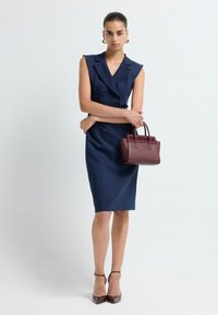 Femme dans une robe blazer sans manches bleu marine, tenant un sac à main bordeaux, portant des chaussures à talons avec bride au cheville bordeaux et des boucles d'oreilles en or, se tenant devant un fond blanc.