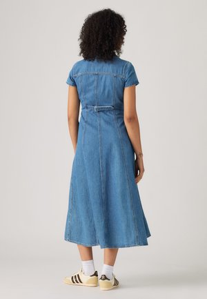 Kvinde med krøllet hår iført en tætsiddende blå denim midi-kjole og hvide sneakers, stående med ryggen til kameraet mod en ensfarvet baggrund.