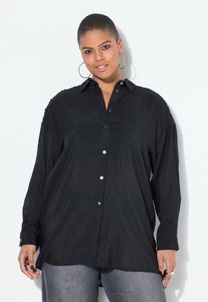 Camisa negra de botones con mangas largas, tejido texturizado, cuello y detalle de bolsillo. Presenta botones brillantes y un corte relajado.