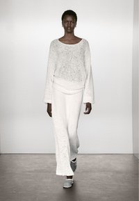 Biały, dziergany sweter z szerokim dekoltem i długimi r�ękawami w zestawie z dopasowaną, długą spódnicą. Modelka ma na sobie szare wsuwane buty.