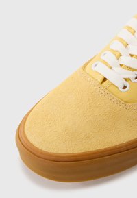 Yellow suede sneaker with white laces, featuring a textured upper and a gum rubber sole. Rounded toe and stitched detailing.
