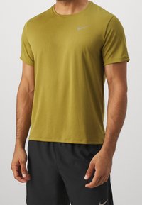 Camiseta atlética de manga corta en verde oliva con cuello redondo y un logotipo reflectante de Nike en plata en el pecho. Tejido de textura suave.