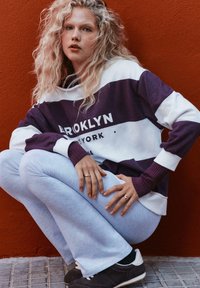 Biely a fialový pruhovaný sveter s nápisom "BROOKLYN", spárovaný so svetlošedými teplákmi a tmavými teniskami, na červenom pozadí.