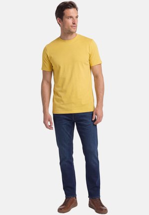 Man draagt een mosterdgele T-shirt met korte mouwen, een blauwe spijkerbroek en bruine sneakers, staand tegen een witte achtergrond, kijkt opzij.