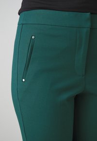 Green pants with a smooth texture, featuring a sleek design, a side pocket with a silver snap, and clean lines along the thigh.