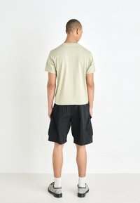 T-shirt vert clair à manches courtes, shorts cargo noirs avec poches latérales, chaussures grises et chaussettes blanches ; fond uni.