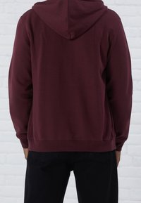 Osoba oblečená v jednobarevné vínové mikině s kapucí staženou a černých kalhotách, stojící proti bílé cihlové zdi, zády k fotoaparátu.