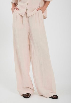 Jasnoróżowe szerokie spodnie wykonane z lekkiego materiału. Projekt charakteryzuje się luźnym krojem i subtelną teksturą. Noszone z czarnymi butami z siateczki.
