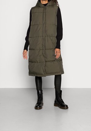 Femme portant une longue veste matelassée vert olive avec capuche, un haut à manches longues noir, un pantalon en cuir noir et des bottes noires à lacets.