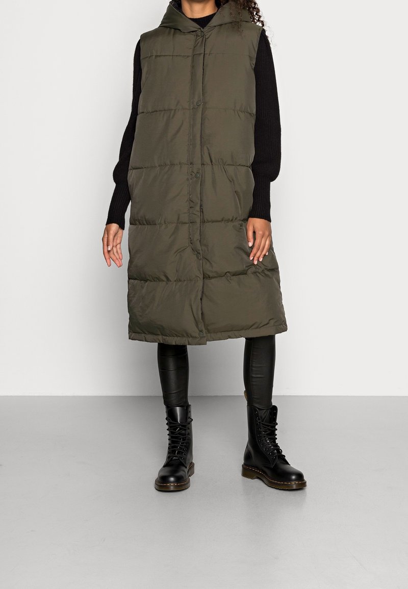 Femme portant une longue veste matelassée vert olive avec capuche, un haut à manches longues noir, un pantalon en cuir noir et des bottes noires à lacets.