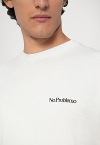 Valge puuvillane T-särk ümmarguse kaelusega ja musta "No Problemo" tekstilogoga ees. Sile tekstuur ja joonis, mis sobib igapäevaselt kandmiseks.