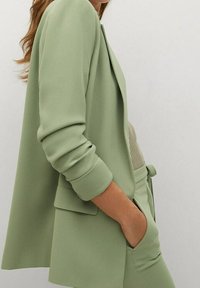 Světlý zelený blazer z hladkého materiálu, s uvolněným střihem, dlouhými rukávy, klopovým límcem a předními kapsami. Detail vymodelovaného střihu.