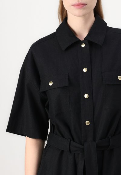 Robe noire à manches courtes avec un col, des boutons-pression dorés, deux poches poitrine et une taille ceinturée pour un ajustement réglable. Fabriquée en tissu texturé.