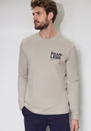 Beige Langarm-T-Shirt aus weichem Baumwollmaterial, mit einem dunkelb blauen "BE FEARLESS" Grafik auf der Vorderseite und einem runden Halsausschnitt.