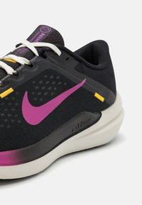 Zapatilla deportiva negra con parte superior de malla texturada, un swoosh de Nike en púrpura, cordones blancos y detalles en amarillo. Suela redondeada con un acabado suave.