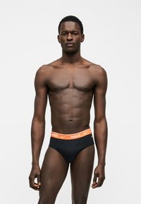 Αθλητικός άνδρας με σκούρο δέρμα, όρθιος μπροστά, φοράει μαύρα briefs με πορτοκαλί μέση Nike σε απλό λευκό φόντο.