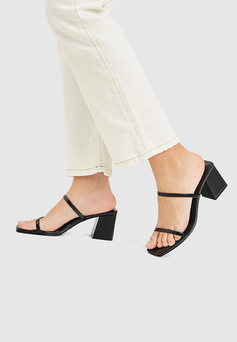 Pieds portant des sandales noires à talons blocs avec brides, associées à un pantalon court écru, sur un fond uni.