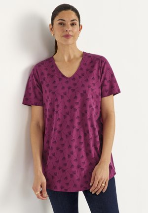 Vrouw met donker haar naar achteren getrokken, draagt een paars V-hals shirt met een zwart bladmotief en donkere spijkerbroek, staand tegen een witte muur.
