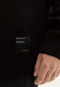 Schwarzer Strickpullover mit strukturiertem Muster, gerippten Bündchen und einem Etikett mit Markendetails, das einen lässigen Stil festlegt.