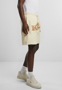 Cream-colored fleece shorts feature an embroidered logo in tan. Elastic waistband with drawstring. Worn with white ankle socks and beige sneakers.