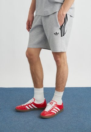 Man die grijze Adidas shorts en rode Adidas sneakers met witte strepen draagt, staat op een blauw tapijt met zijn handen in zijn zakken.