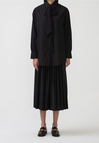 Camicia nera a maniche lunghe con dettaglio della cravatta a farfalla, abbinata a una gonna nera plissettata e mocassini neri con un embellimento dorato.