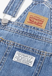 Μπλε τζιν φορμάκι με μεταλλικά κλιπ, χρυστή αντίθεση στο ράψιμο και δύο ετικέτες: μία καφέ με λογότυπο και μία λευκή με κείμενο.