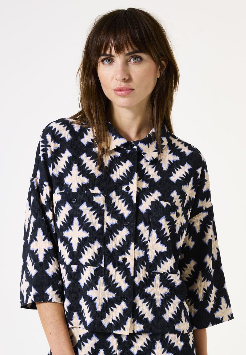 Marineblaues Hemd mit Knopfverschluss aus strukturiertem Stoff, das ein geometrisches Muster in Creme und Hellblau aufweist. Verfügt über zwei Brusttaschen und weite Ärmel.