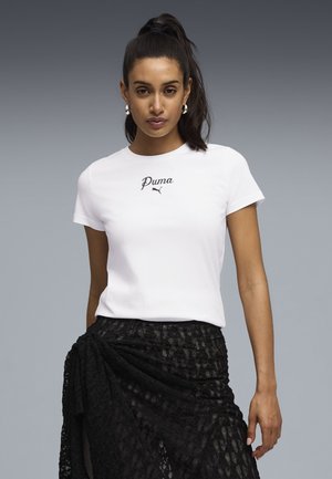 Donna con capelli scuri raccolti in una coda alta che indossa orecchini a cerchio d'argento, una maglietta bianca Puma e una gonna avvolgente di pizzo nera su uno sfondo grigio.