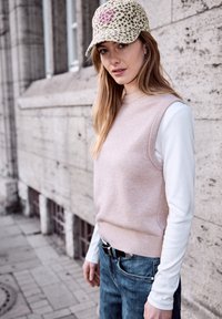 Femme portant une casquette à motif léopard, un pull sans manches beige par-dessus une chemise blanche à manches longues, un jean bleu, debout près d'un mur en pierre en plein air.