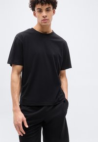 Νέος άνδρας με σγουρά μαλλιά, που φοράει απλό μαύρο κοντομάνικο πουκάμισο και μαύρο παντελόνι, στέκεται μπροστά σε ανοιχτό γκρι φόντο.