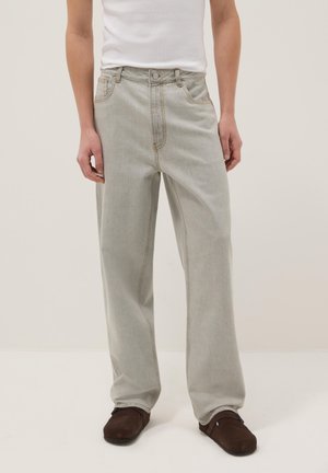 Persona che indossa jeans larghi grigio chiaro, maglietta bianca senza maniche e scarpe slip-on marrone scuro, in piedi contro uno sfondo chiaro e uniforme.