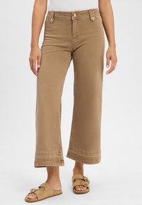 Pantaloni a gamba larga color beige realizzati in cotone, con vita alta, due tasche frontali e dettagli cuciti all'orlo. Abbinati a sabot beige chiaro.