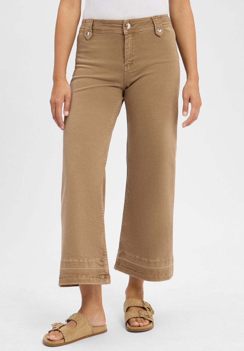 Pantaloni a gamba larga color beige realizzati in cotone, con vita alta, due tasche frontali e dettagli cuciti all'orlo. Abbinati a sabot beige chiaro.
