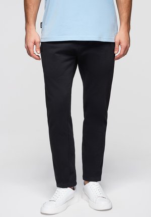 Uomo con pantaloni neri a gamba stretta, sneakers bianche e camicia azzurra, in piedi davanti a uno sfondo grigio uniforme.