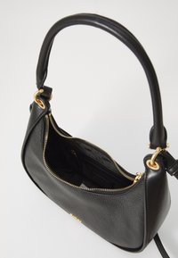 Czarna skórzana torba na ramię z złotym zamkiem i metalowymi elementami, częściowo otwarta, ukazująca czarne materiałowe wnętrze oraz małą kieszonkę na zamek wewnątrz.