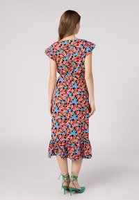 Robe midi florale avec des manches courtes à volants, présentant une base noire avec des motifs rouges, bleus et verts, et un ourlet à volants. Accentuée par des talons verts.