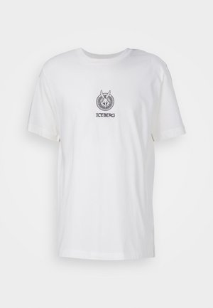Mies, jolla on valkoinen lyhythihainen t-paita, jossa on logo, revityt siniset farkut polvireiällä, mustat kengät ja hopeinen kaulakoru, seisoo käsi taskussa.