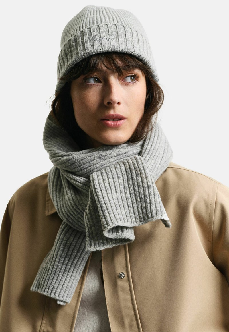 Grå ribbet strikket hue og matching tørklæde, der har en blød tekstur og et enkelt design. Båret over en beige jakke.