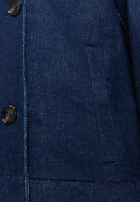 Jeansjacke in Dunkelblau mit Knopfverschluss, strukturierter Stoff und einer Seitentasche als Detail. Nähte sind entlang der Kanten sichtbar.