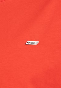 T-shirt rossa realizzata in morbido cotone, con un piccolo etichetta bianca con testo nero "SMILODOX" cucita sul lato. Texture liscia, design aderente.