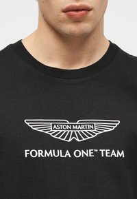 Εικόνα κοντινού πλάνου ενός ατόμου που φοράει μαύρο μπλουζάκι με λογότυπο φτερών της Aston Martin σε λευκό και το κείμενο «FORMULA ONE TEAM» από κάτω.