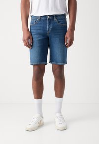 Shorts en denim bleu foncé à coupe droite, dotés de poches avant, d'une taille avec passants de ceinture et d'un ourlet brut. Portés avec des baskets blanches et des chaussettes.