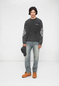Sudadera gris con estampado blanco y detalles desgastados, jeans azul claro y botas de trabajo marrones. Sostiene una gorra gris en una mano.