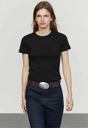 Junge Frau mit langen braunen Haaren trägt ein schwarzes T-Shirt, dunkle Jeans und einen braunen Gürtel mit einer großen ovalen silbernen Schnalle.