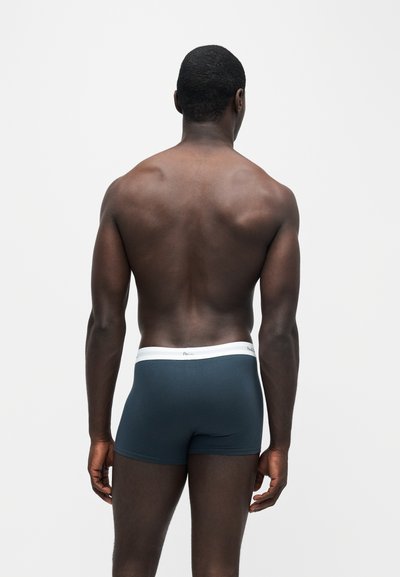 Homme debout, dos à la caméra, portant un boxer bleu foncé avec une ceinture blanche sur un fond uni.