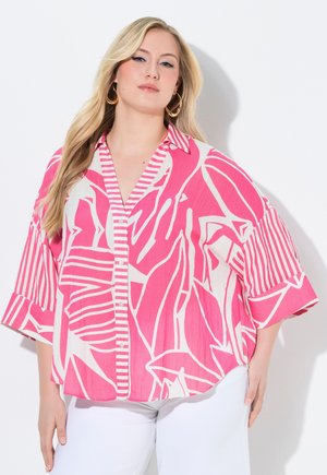 Blonde Frau mit goldenen Creolen und einem hellen, pink-weißen gemusterten, locker sitzenden Hemd mit Knöpfen und weißen Hosen.