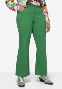Vaqueros verdes acampanados de textura suave, con bolsillos frontales y cierre de botón. Combinados con botas de tobillo metálicas para un contraste.