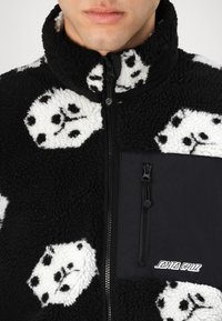 Fleece jacket featuring a black and white dice pattern, high collar, zip closure, and a black fabric chest pocket with logo detail.